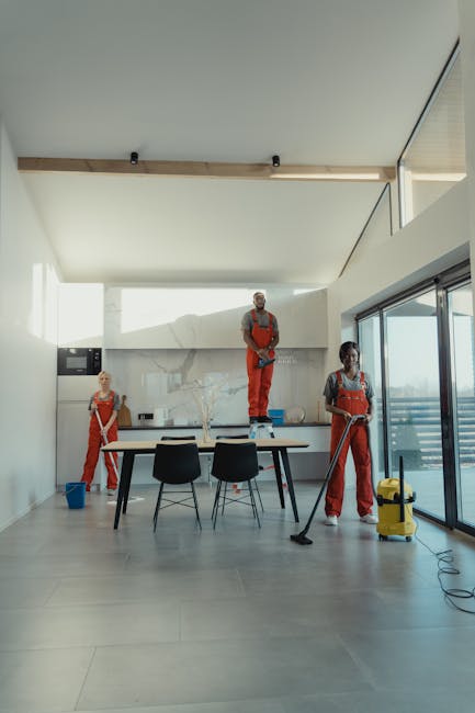 Two professional cleaners from Forestdale Carpet Cleaners are engaged in a domestic surface cleaning task inside a modern living room. One cleaner, a man, is kneeling on the light grey wooden floor, h